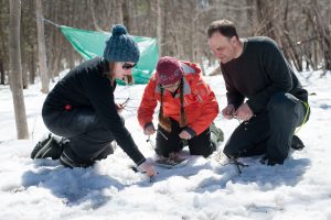 Three people holding twigs starting small fire in the snow